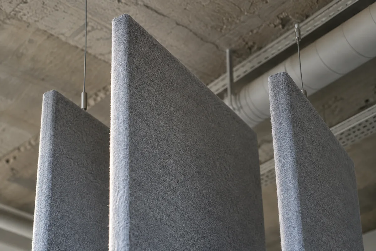 Détail d’un panneau acoustique suspendu en baffle sous un plafond de bureau, matière textile mate, lumière du jour rasante