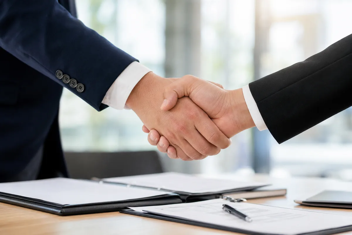 Deux professionnels se serrant la main au-dessus d’une table de réunion avec des dossiers ouverts, signe d’un partenariat conclu